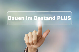 Das Bild zeigt eine Hand, die einen transparenten Text mit der Aufschrift „Bauen im Bestand PLUS“ berührt. Der Hintergrund ist verschwommen.